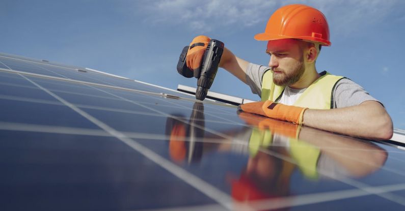 Sustainability Impact - Low angle of serious bearded male electrician in hardhat and protective gloves installing solar photovoltaic panel system using drill