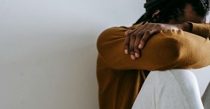 Embracing Failure - Crop anonymous depressed African American male embracing knees and covering face with arms while sitting on floor against white wall
