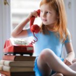 Small Talk - Low angle of calm redhead preteen lady in blue dress and beige sandals looking away and having phone call using retro disk telephone on stack of books while sitting with legs crossed on wooden table against window at home